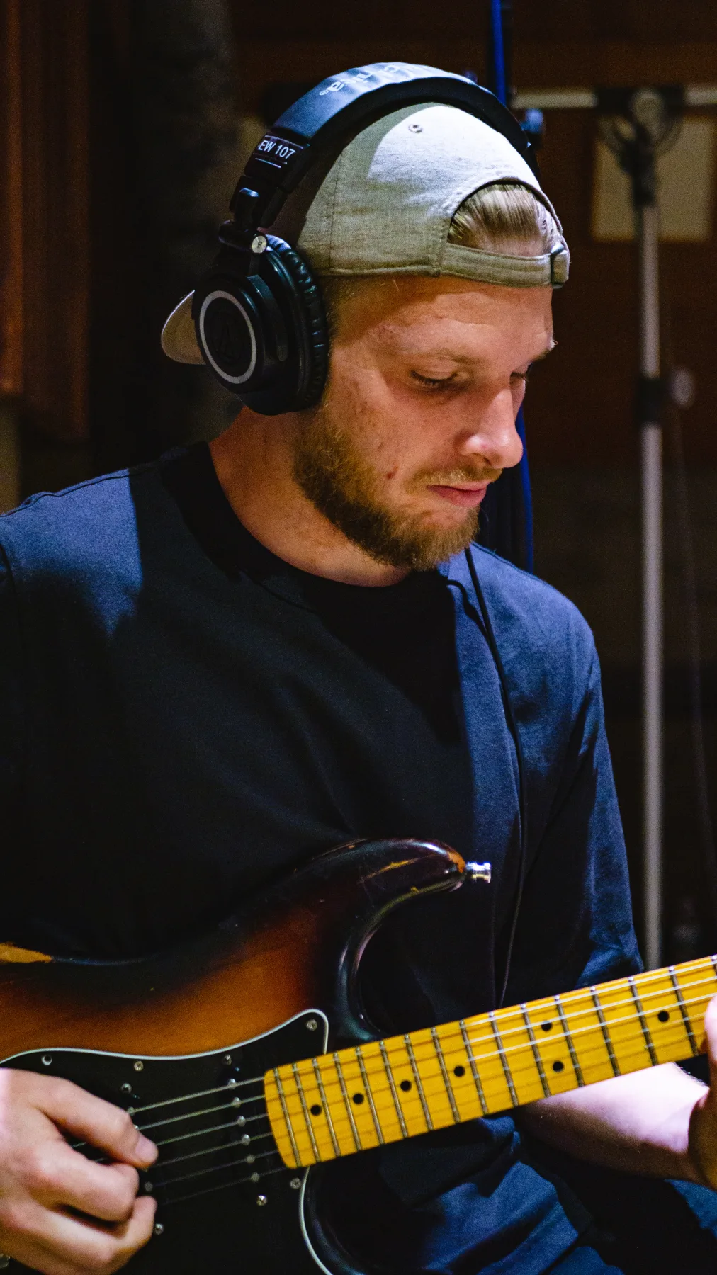 Musician wearing headphones and a backward cap while playing bass in a recording room.