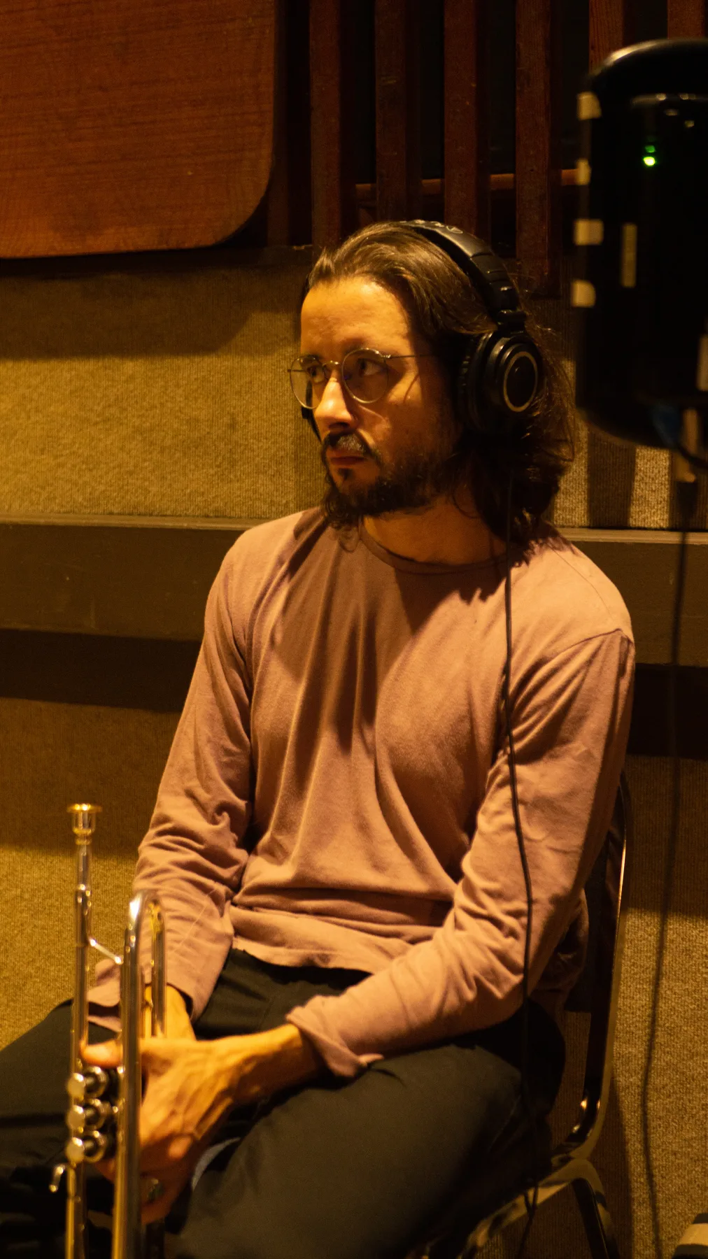 Musician wearing headphones and holding a trumpet in a warmly lit recording room.