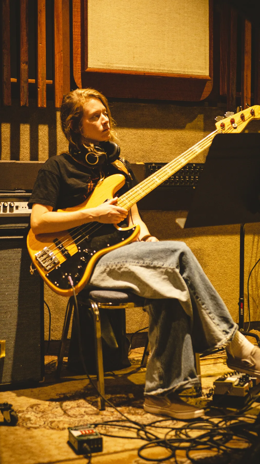 Musician with headphones seated on a stool while holding an electric bass in a recording room.