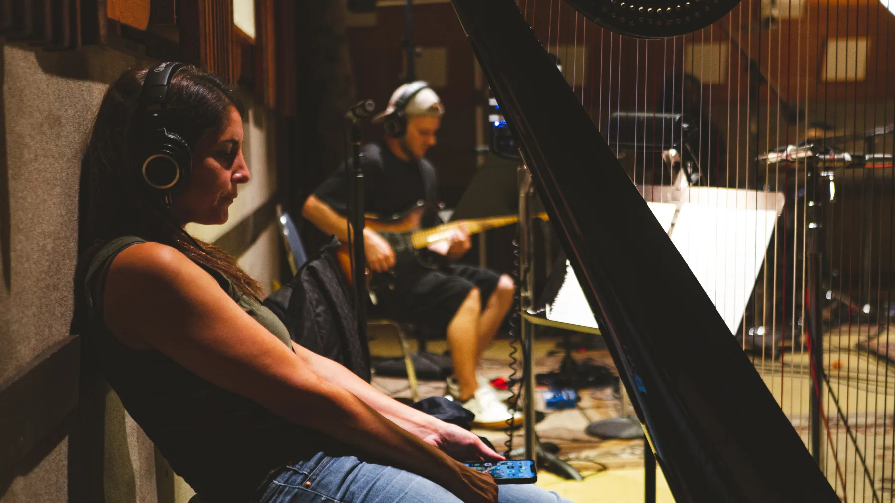 Musician wearing headphones seated beside a harp while another musician plays bass in a recording room.